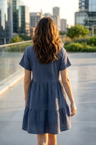 Cassady Dress featuring a loose, comfortable silhouette with a V-neckline, short sleeves, and tiered skirt, shown from the back on a rooftop walkway with greenery and city buildings in the distance.