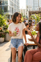 Strawberry Coquette American Vintage Graphic Crop Top featuring a soft cream fabric with playful strawberry coquette print, styled with denim shorts and shown in a lively outdoor café setting.
