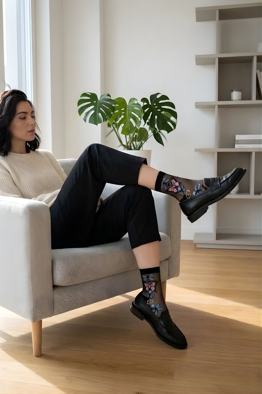 Butterfly Sheer Socks 2-Pack featuring transparent mesh fabric with intricate butterfly designs, styled in black paired with loafers while seated in a minimalist living room.