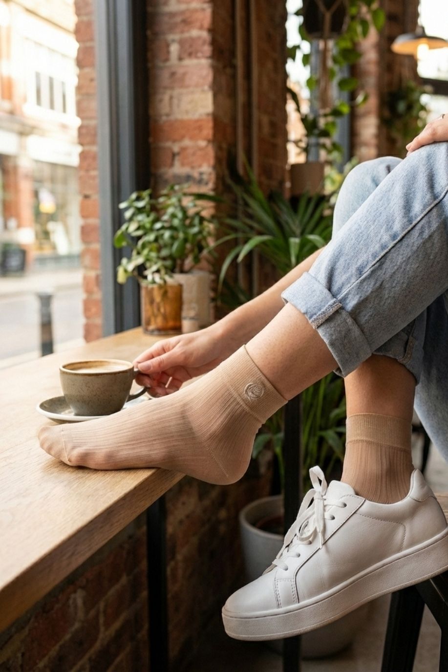 Beige sheer ribbed ankle socks with subtle rose embroidery styled with white sneakers and cuffed jeans, shown in a cozy café setting with a coffee cup.