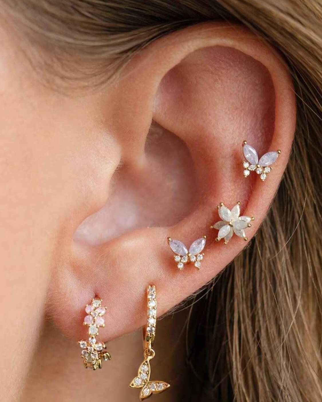 Remember Me Butterfly Studs featuring marquise cut butterfly crystals with shimmering stone accents and a gold tone setting, styled in a curated ear stack with matching floral and hoop earrings, emphasizing layered jewelry styling and sparkling detail.