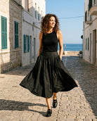 Poplin Cotton Smocked Flare Midi Skirt featuring a full midi shape with gathered volume, structured waistband, and practical side pockets, worn while walking through a sunlit seaside street, highlighting movement and relaxed styling.
