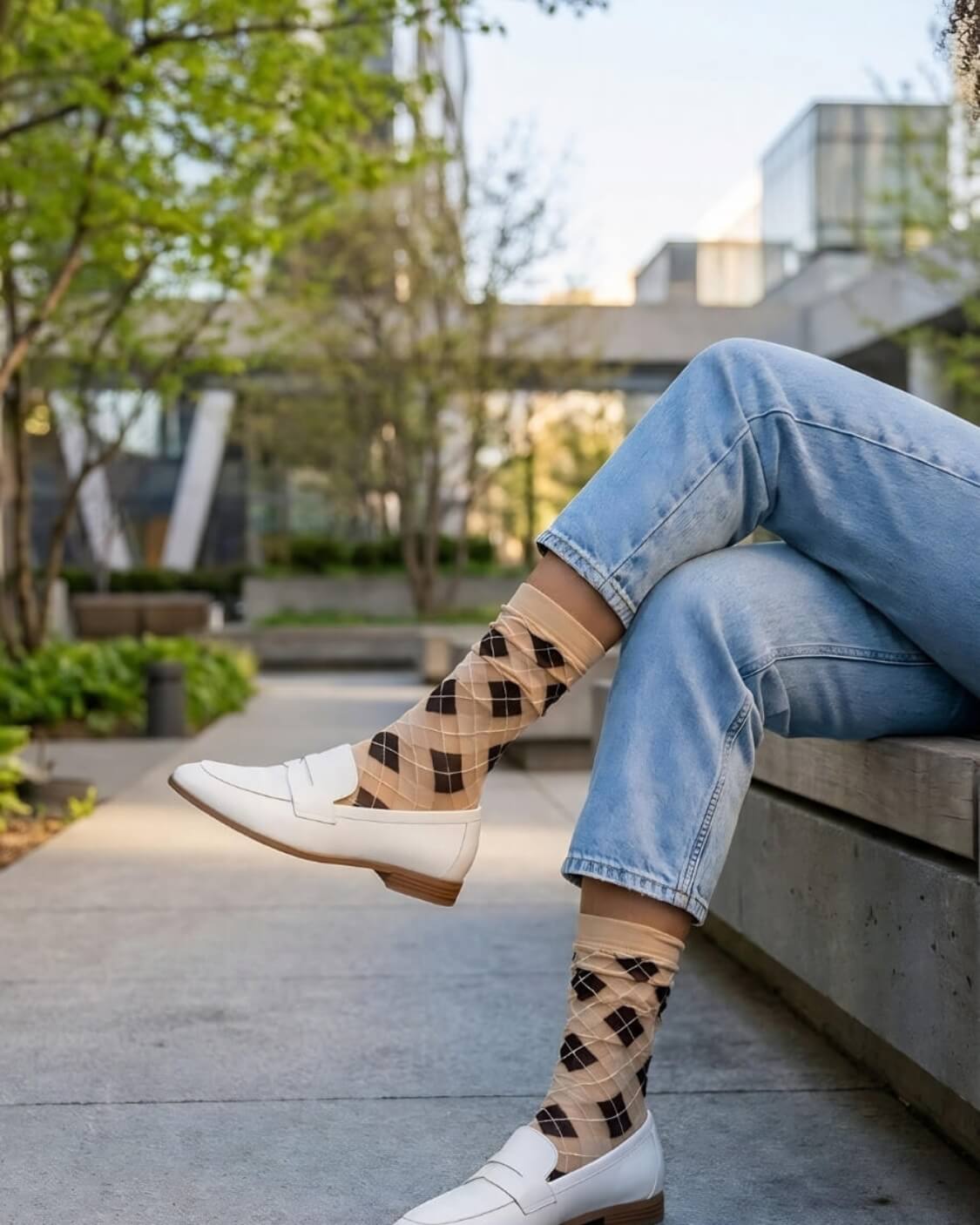 Sheer Argyle Crew Socks in beige and black featuring a soft diamond pattern, paired with white loafers and cuffed jeans while sitting on a concrete bench, showcasing the lightweight, sheer texture and casual styling.