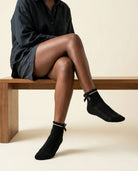 Black ribbed ankle socks with scalloped cuff and black satin bow, legs crossed on a wooden bench