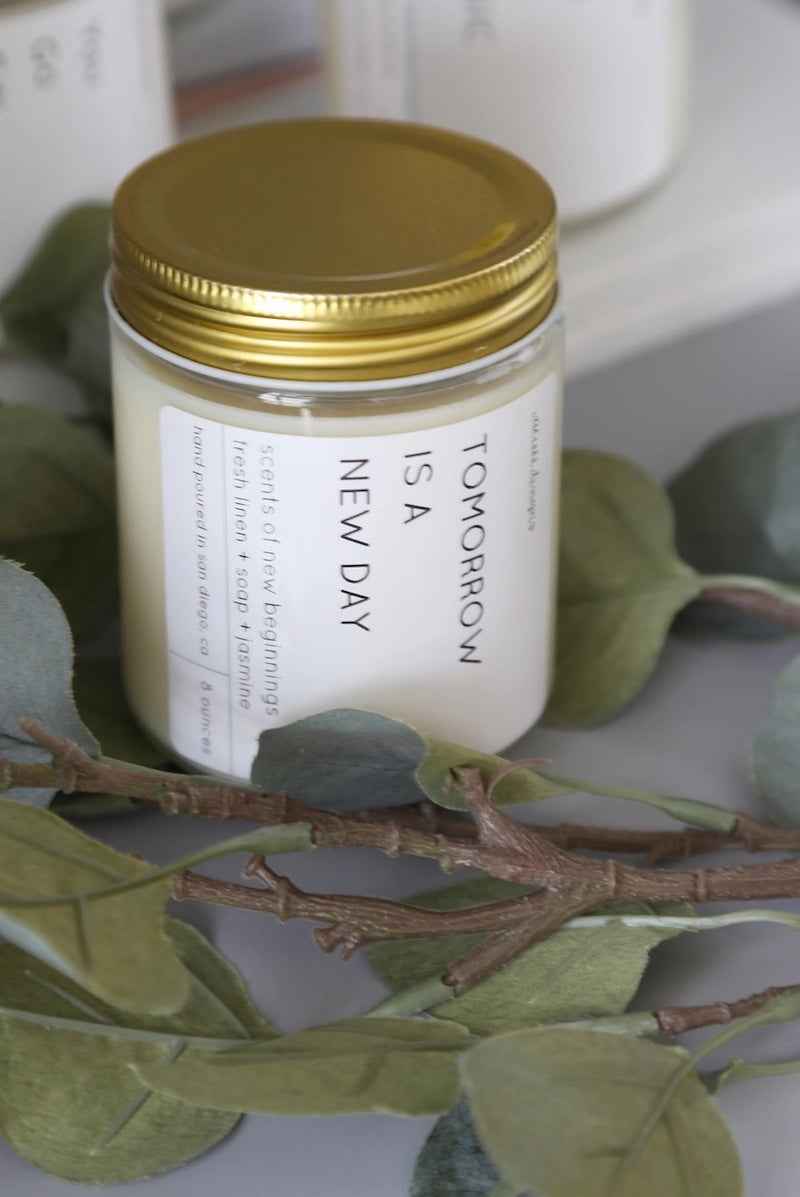 Love Classic hand-poured candle with gold lid and 'Tomorrow is a New Day' label, styled with greenery.