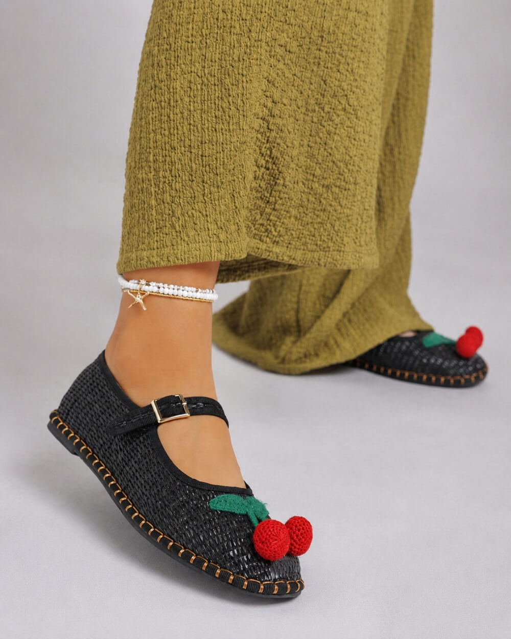 Cherry Accent Mary Jane Flats, black woven Mary Jane flats with cherry detail and buckle strap, paired with a textured olive-green dress and minimal anklet, showcasing a relaxed yet playful daytime look.
