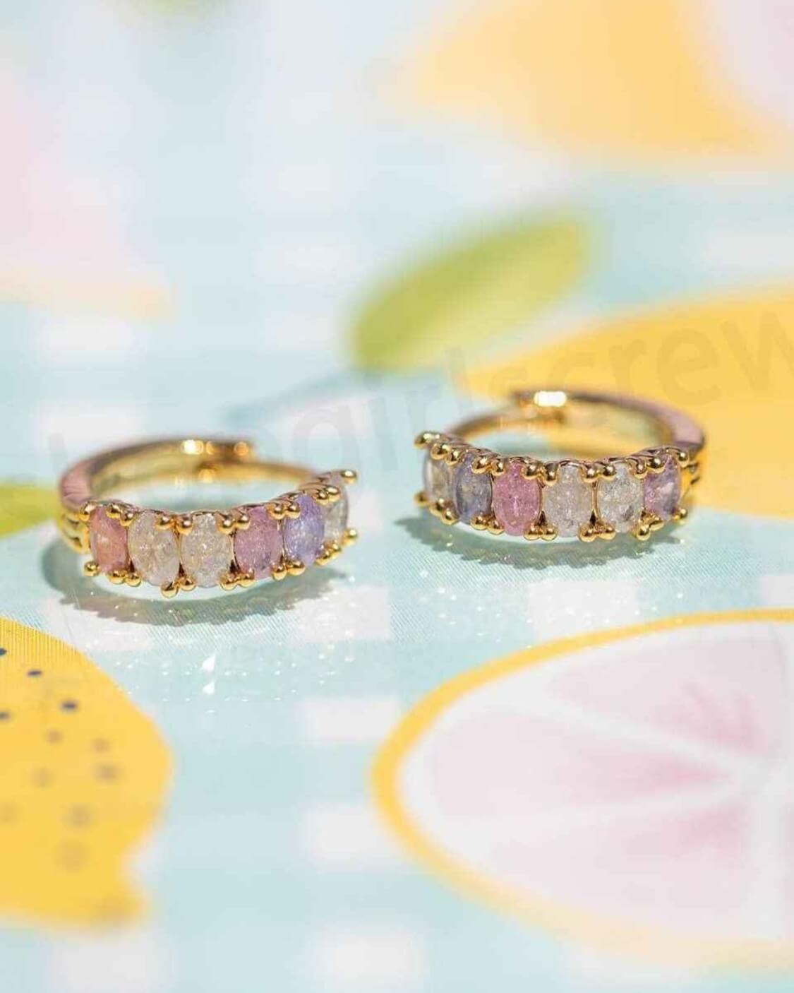 Dreaming About You Hoops featuring pastel oval stones set along a polished gold tone hoop silhouette, displayed in a close up angled product shot on a colorful printed backdrop, highlighting soft sparkle and refined curved structure.