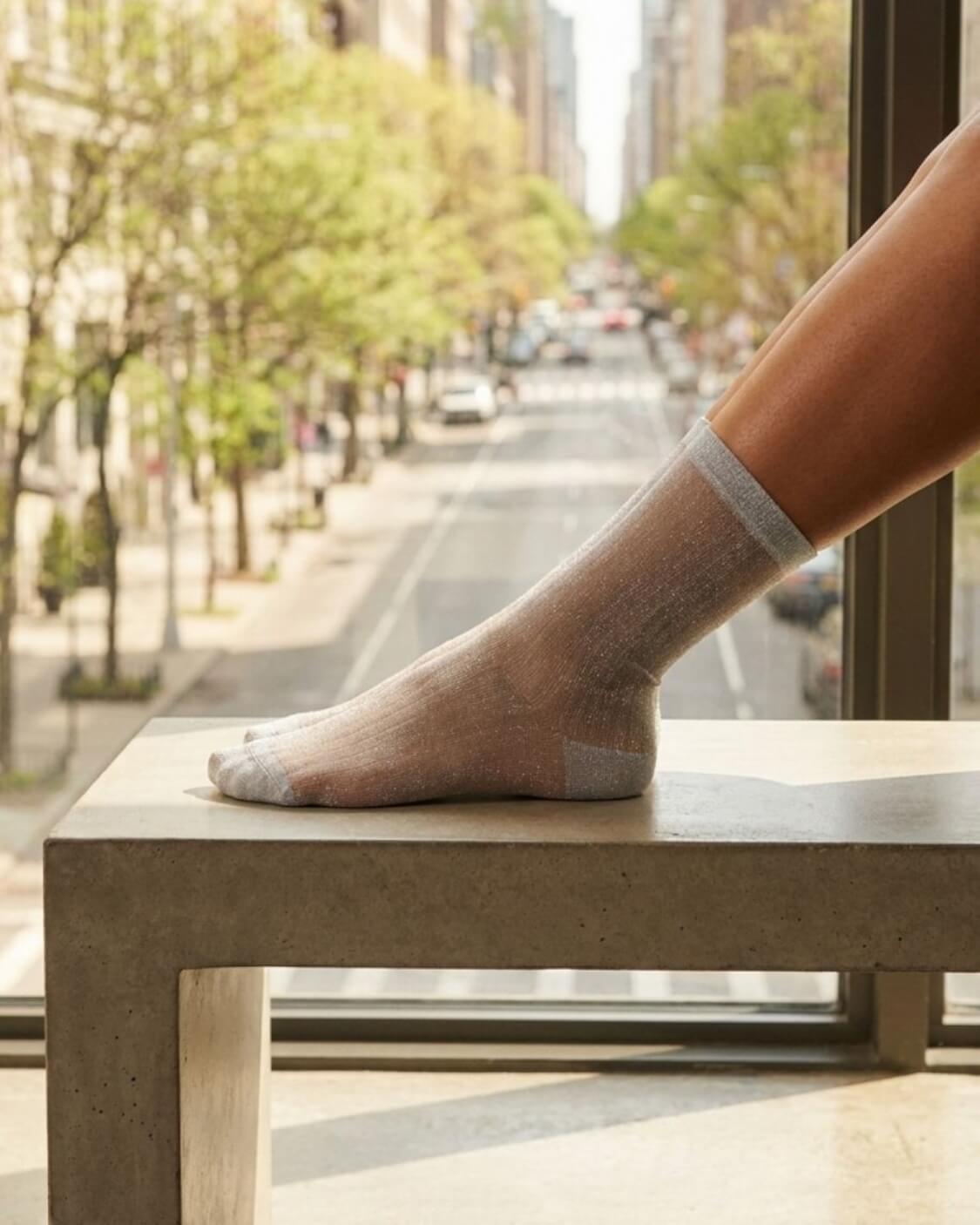 Sheer Metallic Ribbed Crew Socks in sheer silver tone with metallic sheen, displayed on a foot resting on a concrete ledge by a city window, showcasing the transparent lightweight fabric and smooth ribbed construction.
