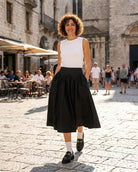Poplin Cotton Smocked Flare Midi Skirt featuring a high waist, gathered flare shape, midi length, and side pockets, styled with a sleeveless top, socks, and loafers in a busy stone plaza, highlighting clean everyday styling.