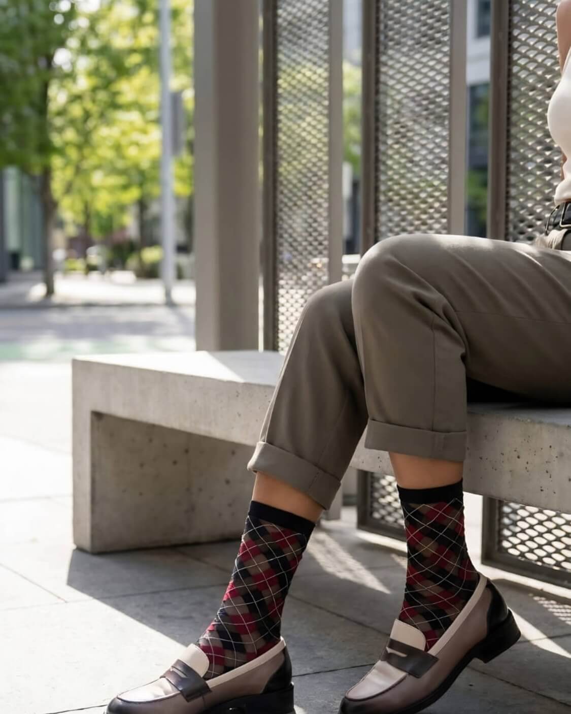 Sheer Argyle Crew Socks in black, red, and beige featuring a classic argyle motif, styled with rolled cuff pants while seated on a sunlit bench, highlighting the semi-sheer material and stylish patterned look.
