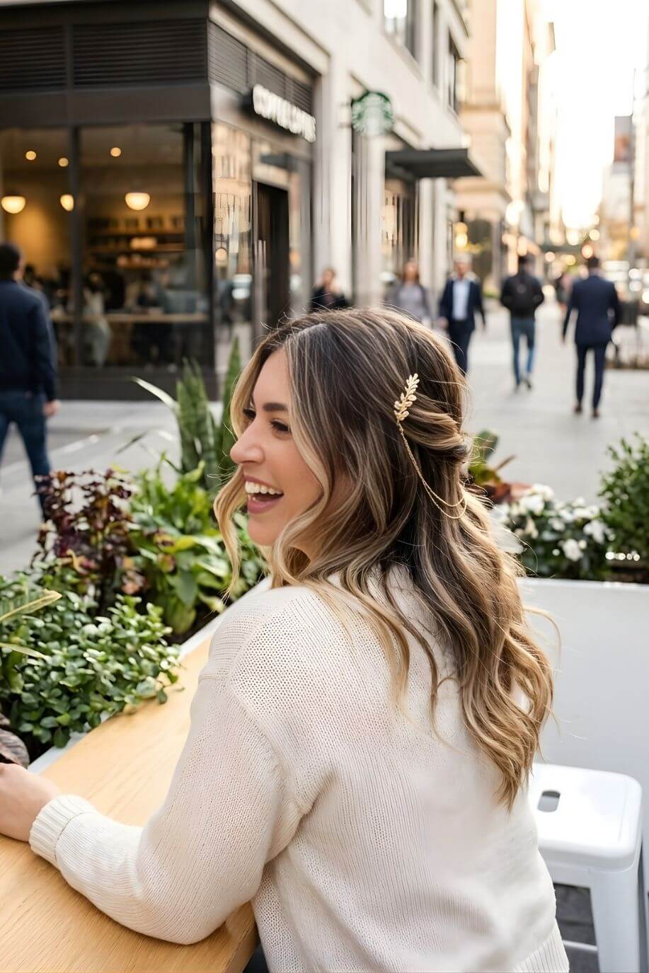 Bloom Bouquets Hair Chain featuring an elegant gold leaf hair clip with fine draped chains, worn on shoulder-length wavy hair, shown in a candid outdoor city café scene with soft sunlight.