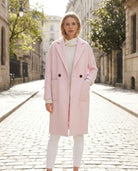 Publisher Double-Breasted Wool Blend Coat in soft blush pink, featuring a relaxed longline silhouette, notched lapel collar, double-breasted button detailing, and oversized patch pockets, styled over a cream knit sweater and white pants while standing on a sunlit cobblestone city street.