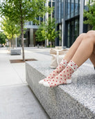 Blushing Hearts Sheer Socks featuring sheer ankle socks with raised red heart accents, soft cuff and toe panels, worn while seated on a stone bench in a bright city setting, highlighting the translucent finish and bold heart pattern.