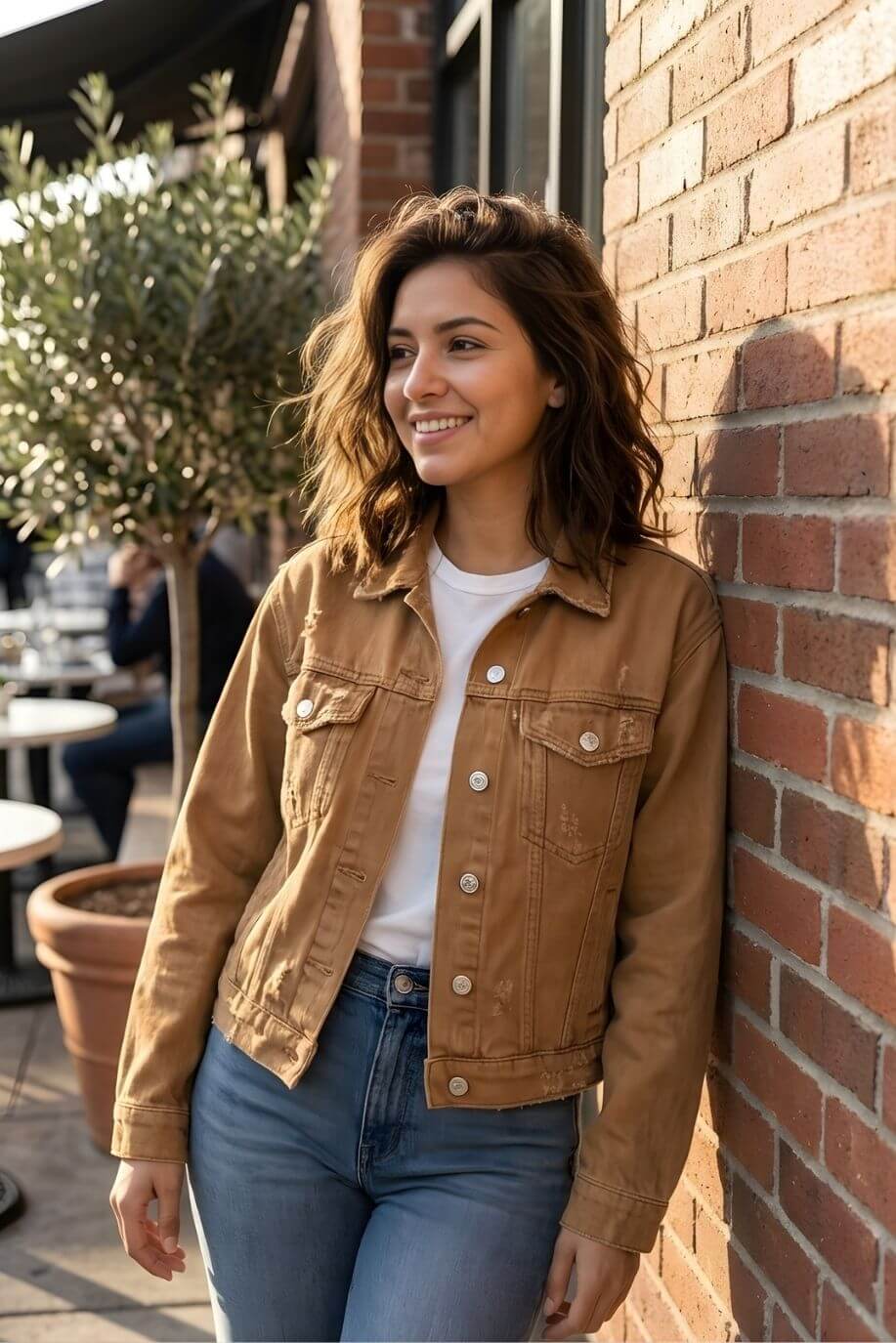 Long Sleeve Denim Jacket in tan, featuring a classic button-front design with chest pockets and slightly fitted silhouette, paired with a white tee and blue jeans, shown on a model leaning against a brick wall in warm natural light.