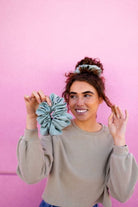 Model wearing the Eucalyptus Jumbo Scrunchie against a pink background, featuring oversized sateen fabric and soft eucalyptus green color.