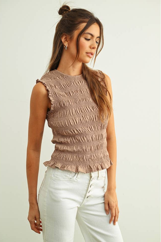 Model wearing the sleeveless smocked top in a neutral tone paired with light denim, highlighting the fitted shape, soft stretch texture, and ruffled hem.