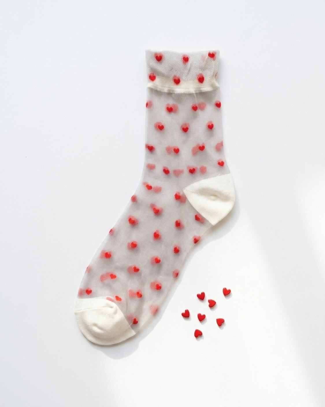 Blushing Hearts Sheer Socks featuring a sheer ankle sock with raised red heart accents, soft cuff, toe and heel panels, displayed flat on a clean studio background with small heart props, highlighting the playful heart texture and lightweight finish.