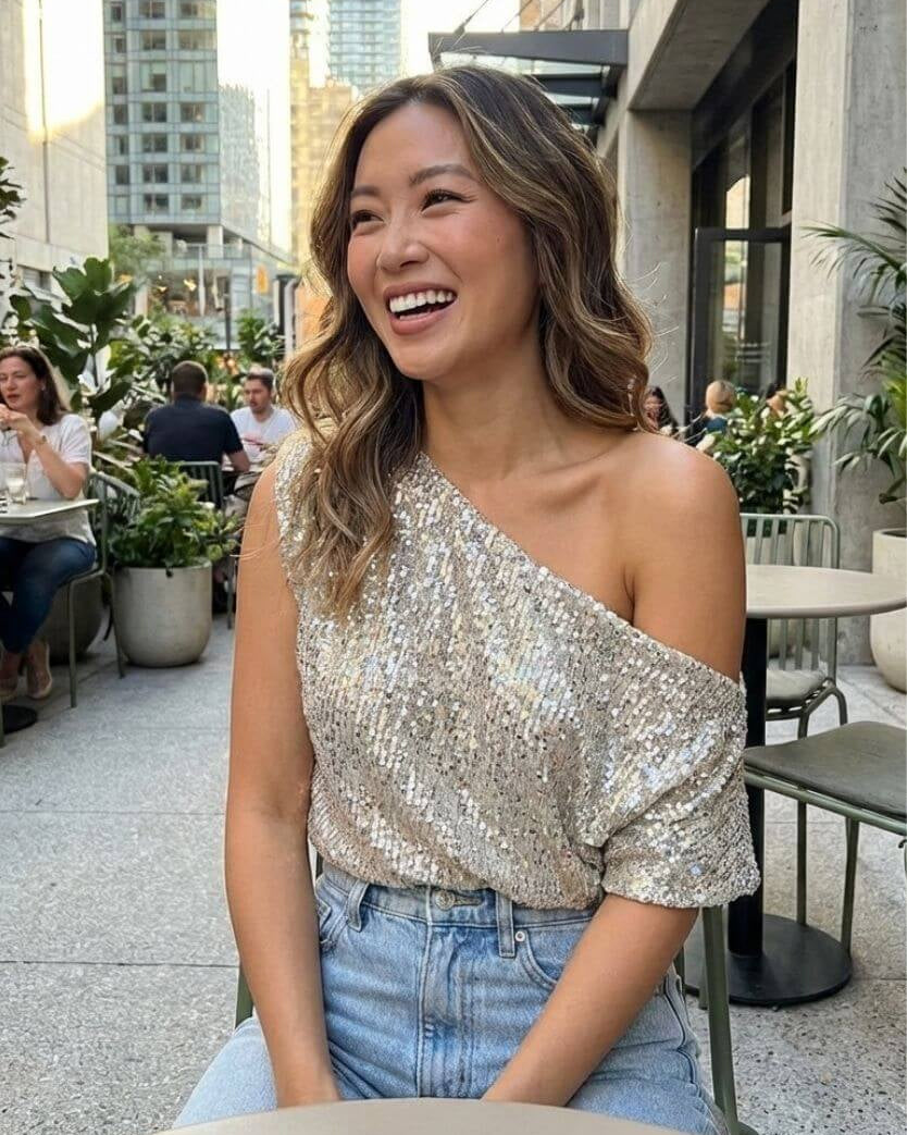 Premiere Night Top one shoulder sequin top styled at an outdoor dining setting with urban background, paired with high waist jeans, highlighting the soft drape and easy going evening style.