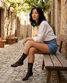 Floral Sheer Socks in Black featuring a transparent floral pattern, ankle-height fit, and smooth top band, worn while seated on a wooden bench in a stone alley, highlighting relaxed styling with delicate texture.