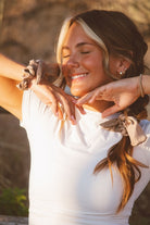 Model wearing the Chestnut Bow Scrunchie styled in a centipede hairstyle and wrapped on the wrist, featuring creamy brown fabric with a soft glossy sheen and bow detail.