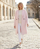 Publisher Double-Breasted Wool Blend Coat in blush pink with a long relaxed fit, notched lapel collar, double-breasted button accents, and oversized patch pockets, worn with a cream sweater, white pants, and sneakers while walking along a charming cobblestone street in daylight.
