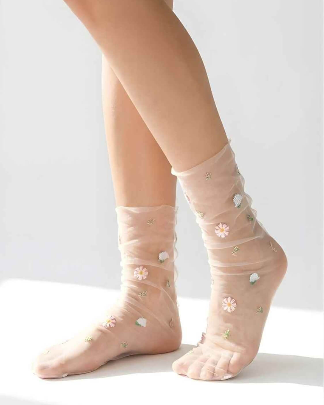 Sheer Garden Daisy Crew Socks in blush nude mesh with embroidered daisy flowers, shown on crossed legs against a clean light background to highlight the transparent fabric.