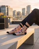 Roses Sheer Socks 2-Pack featuring sheer ankle socks with embroidered rose motifs, black cuff and toe panels, worn while seated on an outdoor bench in a rooftop city setting, highlighting the floral texture and lightweight translucent finish.