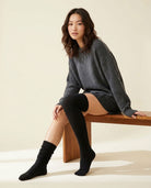 Braided Long Socks in black, one over the knee and one scrunched, with a charcoal sweater on a wooden bench.