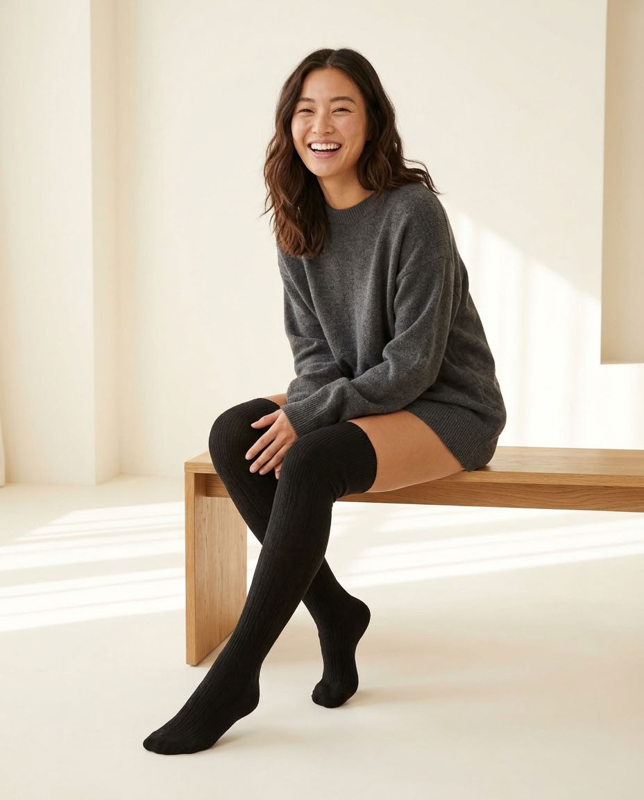 Black Braided Long Socks pulled over the knee, gray sweater, seated on a light wood bench.