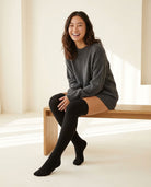Black Braided Long Socks pulled over the knee, gray sweater, seated on a light wood bench.
