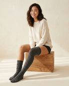 Long Polka Dot Socks in gray with white polka dots, one pulled over the knee and one scrunched at the ankle, styled with a cream sweater on a wooden box.